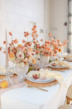 the table is set with peach colored flowers and white plates, silverware, and candles