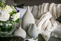 vases and flowers on a glass table in front of a couch with a blanket