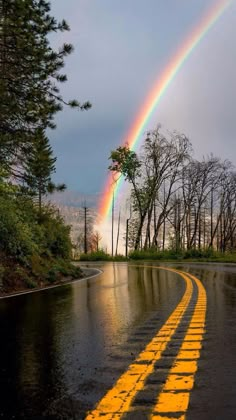 a rainbow appears over a wet road in the middle of the day with trees on either side