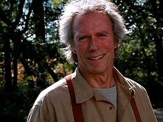 an older man wearing suspenders and smiling at the camera with trees in the background