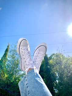 a person with their feet up in the air wearing white converses and jeans, against a sunny blue sky