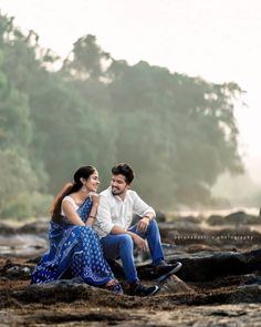 a man and woman sitting next to each other on some rocks near the water with trees in the background