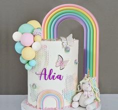 a birthday cake decorated with rainbows, balloons and a unicorn figurine next to it