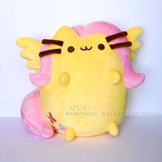 a yellow stuffed animal with pink cheeks and wings on it's back, sitting in front of a white wall