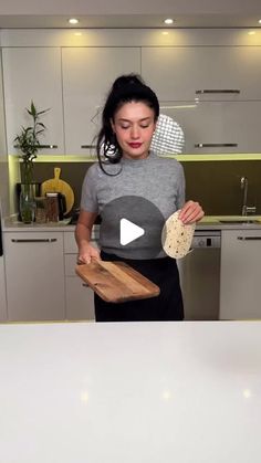 a woman standing in front of a kitchen counter holding a wooden cutting board and bread