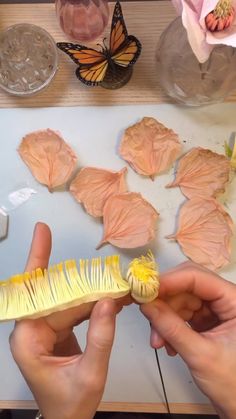 two hands are holding a yellow brush near some flowers and other things on the table