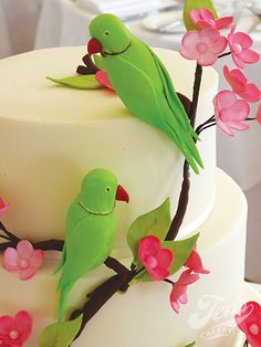 two green birds sitting on top of a white cake with pink flowers and leaves around it