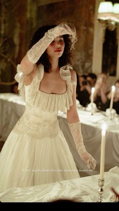 a woman in a white dress standing next to a table with two candles on it