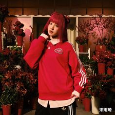 a woman with red hair wearing a red sweatshirt and black pants standing in front of potted plants