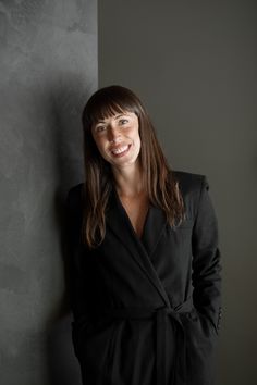 a woman in a black suit standing next to a gray wall with her hands on her hips