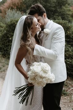 a bride and groom standing together in front of some bushes with white flowers on their bouquets