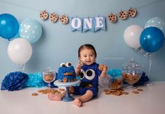 a baby boy sitting on the floor in front of a cake and cookies for his first birthday