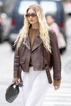 a woman with long blonde hair wearing a brown leather jacket and white pants is walking down the street