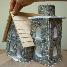 a hand holding a model of a house made out of rocks and wood shingles