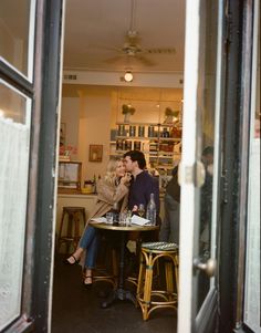 a man and woman sitting at a table in front of a store window looking into each other's eyes