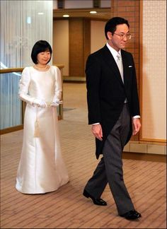 a man and woman dressed in formal wear walking down a hall way, one wearing a long white dress