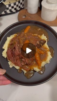 a person holding a black plate with meat and vegetables on top of mashed potatoes