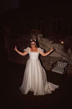 a woman in a white dress and sunglasses posing for the camera at night with her arms outstretched