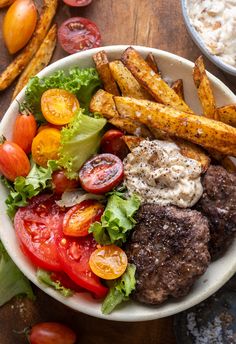 a white bowl filled with meat and vegetables next to french fries on top of a wooden table