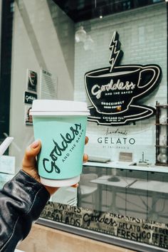 a person holding up a cup of coffee in front of a building with a sign on it