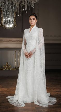 a woman in a white gown and cape standing on a wooden floor with chandelier