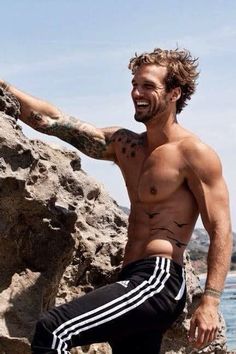 a shirtless man sitting on top of a rock next to the ocean and smiling