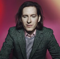 a man with long hair wearing a green jacket and pink shirt sitting in front of a red background