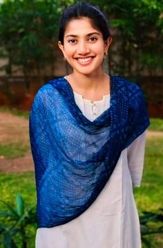 a woman wearing a blue shawl standing in front of some trees and grass, smiling at the camera