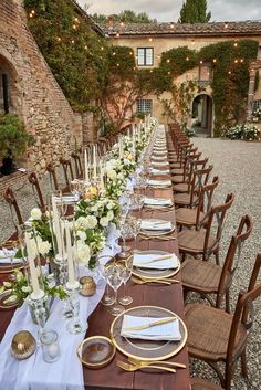 a long table is set with white and gold place settings for an outdoor dinner party