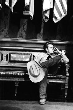 a man sitting on a bench wearing a cowboy hat and drinking from a cup in front of american flags