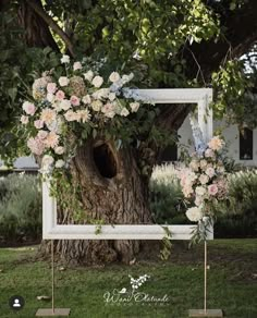 an outdoor ceremony with flowers and greenery on the tree trunk, in front of a white frame