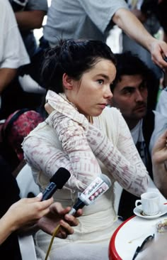 a woman sitting at a table with microphones in front of her and people around her