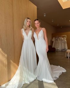 two women standing next to each other in front of a wooden wall wearing wedding dresses