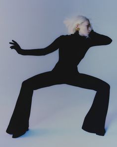 a woman with white hair and black pants is posing in the air while holding her arms out