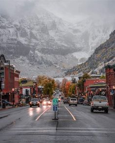 cars are driving down the road in front of snow covered mountains and buildings on either side