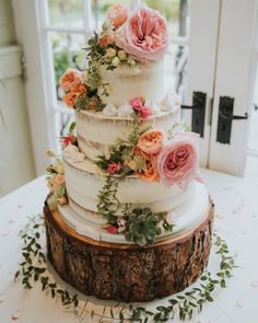 a white wedding cake with pink flowers and greenery on the top is sitting on a tree stump