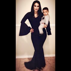 a woman in a black dress holding a baby wearing a long sleeved gown with bell sleeves