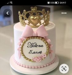 a white cake with pink and gold decorations on top, decorated with a crown for someone's special occasion