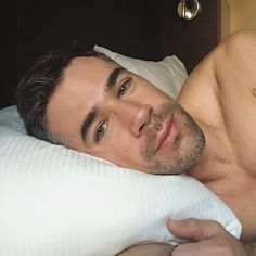 a shirtless man laying in bed with his hand on the pillow and looking at the camera