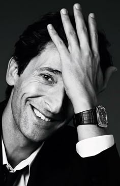 a black and white photo of a smiling man with his hand on his head, wearing a watch