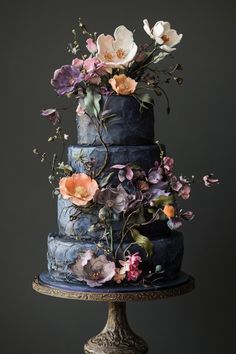 a three tiered blue cake with flowers on the top and bottom, sitting on a pedestal