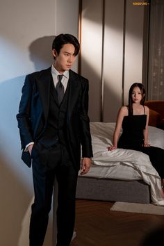 a man in a suit and tie standing next to a woman sitting on a bed