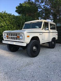 an old white truck parked in front of some bushes and trees on a gravel lot