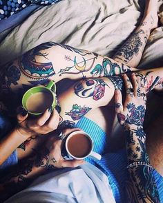 two people laying in bed with tattoos on their bodies and holding coffee mugs next to each other