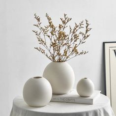 three white vases sitting on top of a table next to a book and plant