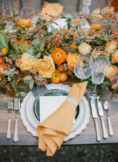 the table is set with silverware, orange flowers and greenery on top of it