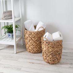 two wicker baskets with toilet paper on the floor next to a white shelf and potted plant