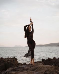 a woman is standing on rocks by the water with her arms up in the air