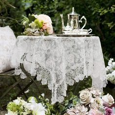 the table is covered with white lace and has flowers on it next to a tea pot