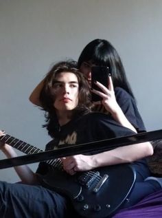 two people sitting on a bed with one holding a guitar and the other using a cell phone